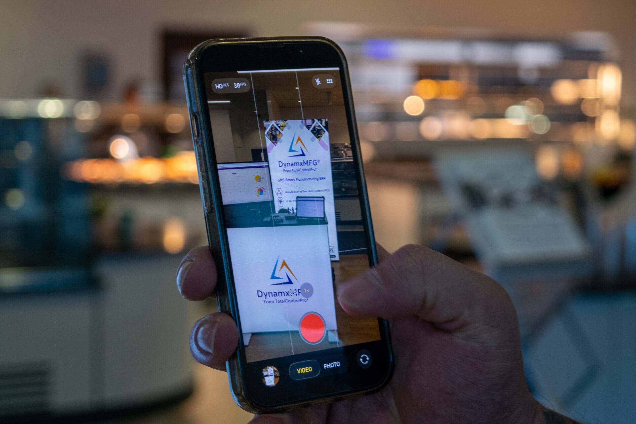 Person photographing DynamxMFG display with smartphone.